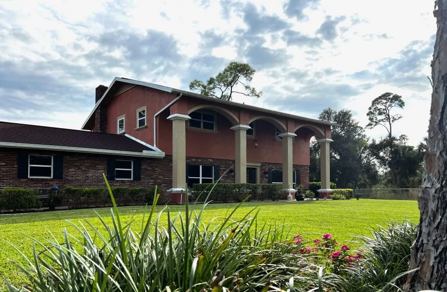 A spacious two-story house with red walls and white columns, surrounded by lush green grass and colorful flowers under a cloudy sky